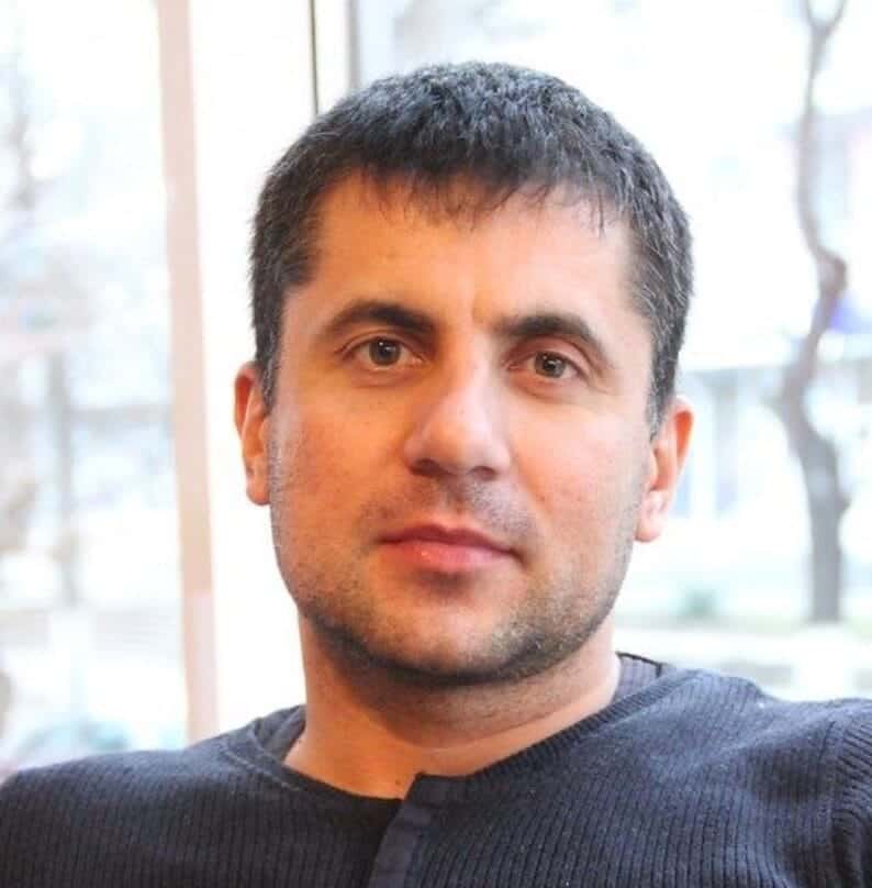 Чоловік із темним волоссям і світлою шкірою, посміхається, сидить у кімнаті біля вікна.